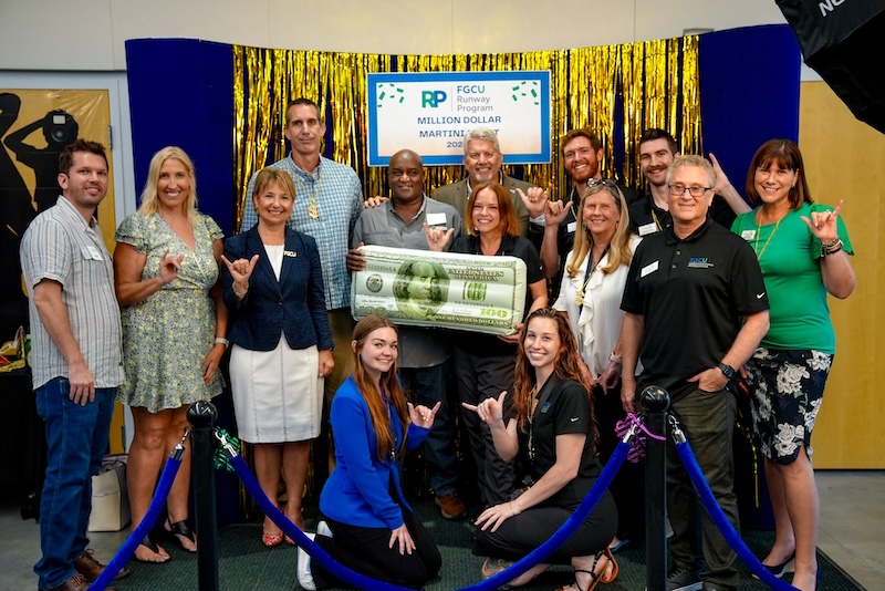 FGCU faculty and staff celebrate Million Dollar Martini Night. Dressed in casual clothing, they are posing behind a gold and blue backdrop as they do the “wings up” sign and hold an inflatable $100 bill. Blue Stanchion ropes can be seen in front of the group.