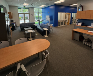 The Military & Veterans Success office lounge features a couch area in the middle of the lounge, a long table with chairs towards the front of the office and a reception desk with cabinets on the right side. Cabinets can also be seen on the left side against the wall. Offices can be seen to the right of the lounge area featuring a blue wall. The back of the lounge area has floor-to-ceiling windows and exit doors.  