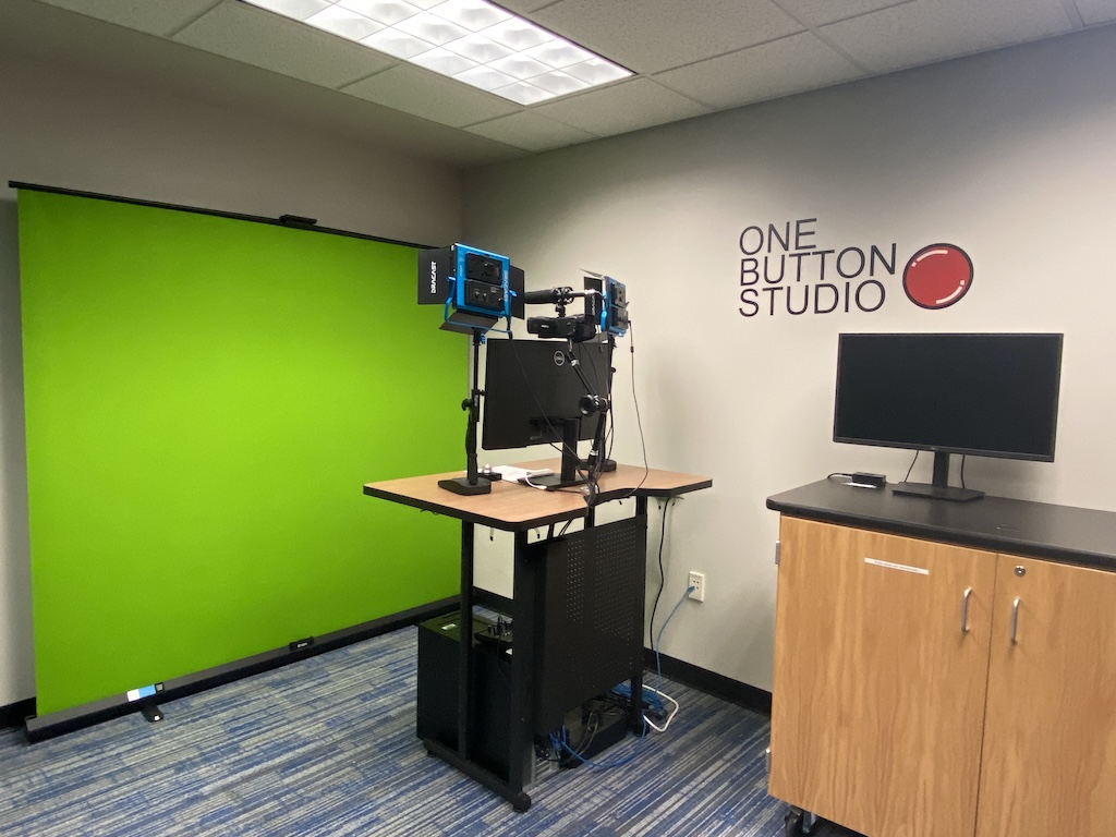 A recording studio setup featuring a green screen backdrop, a desk equipped with mounted cameras and monitors and an additional monitor placed on a cabinet. The wall displays the text 'ONE BUTTON STUDIO' accompanied by a red button graphic.  