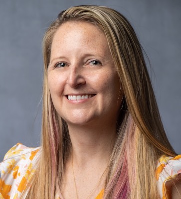 A professional portrait shows a person with long hair wearing a white top with yellow-orange floral accents. The image is framed from the shoulders up against a gray background. 