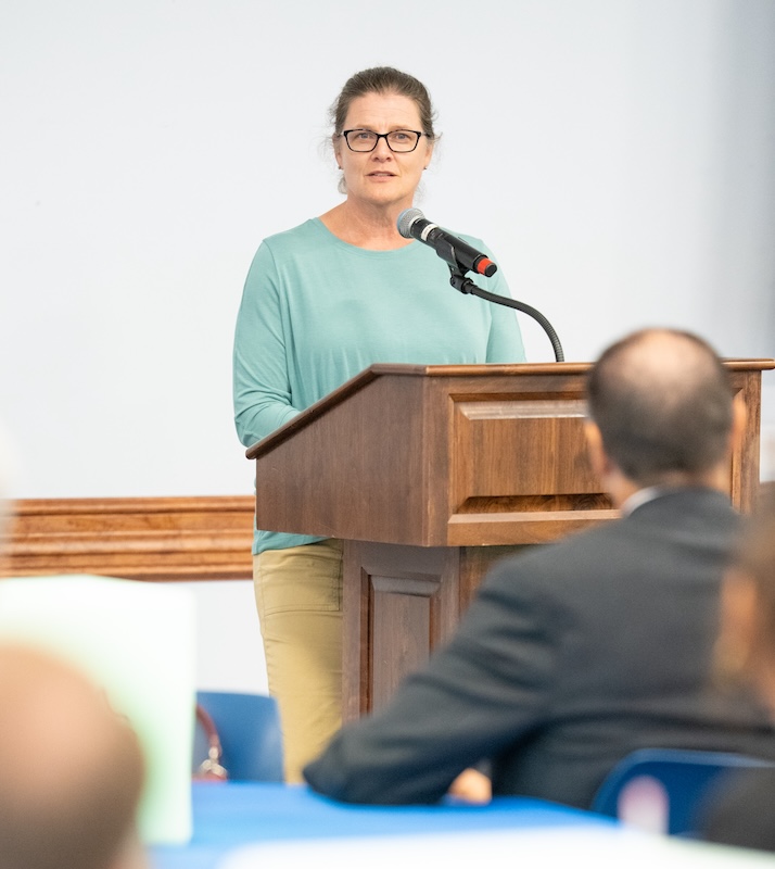 A person wearing glasses, a light teal long‑sleeve shirt and tan pants, stands at a wooden podium speaking into a microphone during an indoor event. Guests seated at tables listen from the foreground.