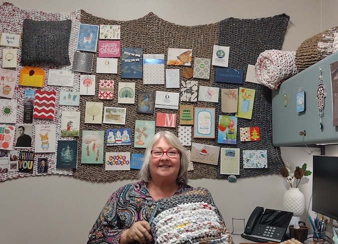 Patty Krupp, wearing red glasses and a colorful blouse with black, blue, pink and orange colors, is displaying plarn creations in her office space. She is holding a white and gray mat made from plastic yarn. On the wall behind her, a white, purple, brown and gray mat is hanging across the wall with ‘happy birthday’ cards pinned to it. Gray overhead storage cabinets are seen on the right side of her office as well as office supplies including two computer monitors, an office phone, a keyboard and pens.