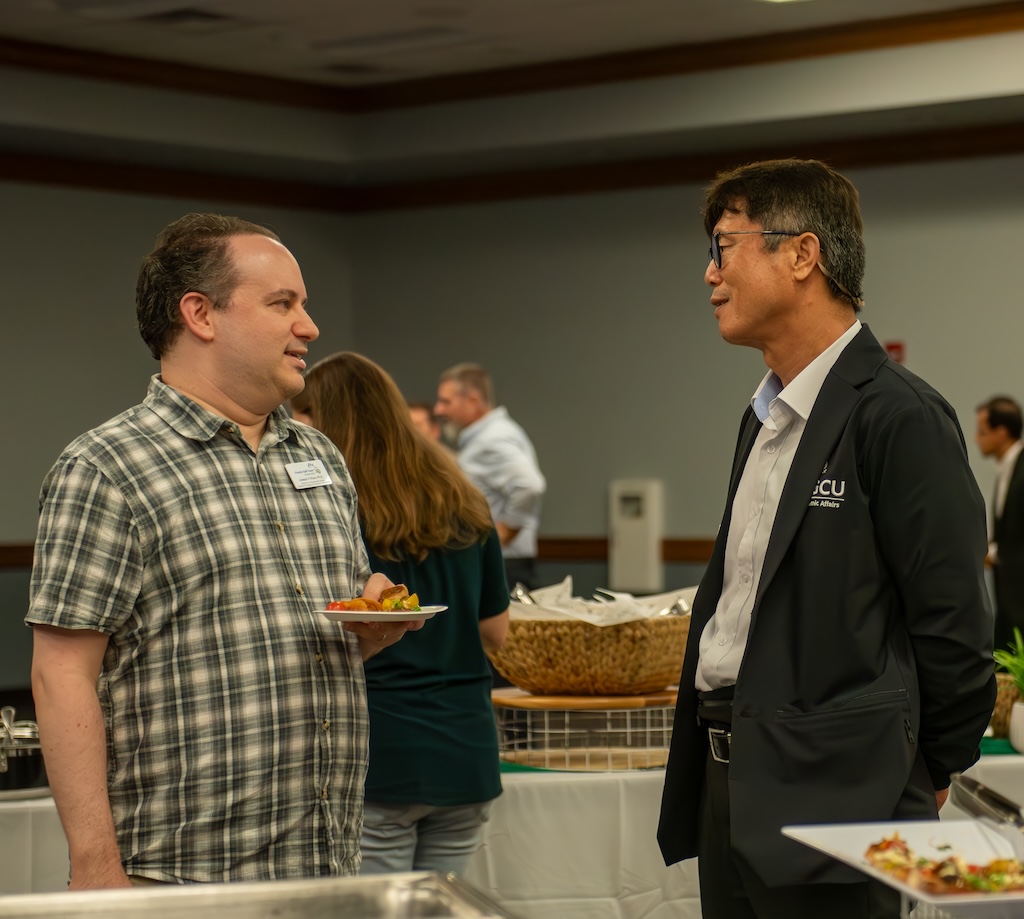 Two individuals are engaged in conversation. One person is holding a plate of food. The person on the left is wearing a short-sleeved, plaid button-up shirt with a name tag on the chest. The other person, on the right, is dressed in a white dress shirt, black trousers, and a dark blazer embroidered with 'FGCU' text. In the background, other attendees and tables with food items are visible. 