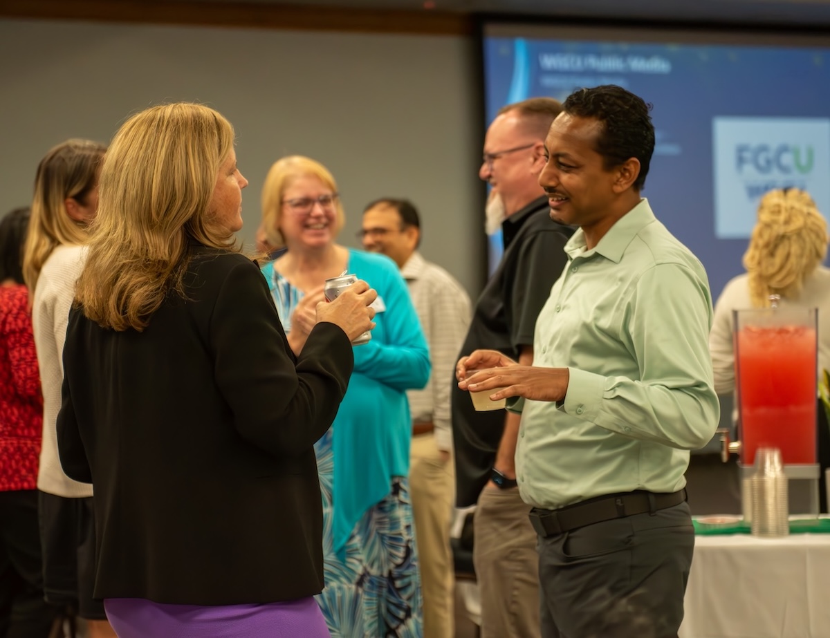 Two individuals are engaged in conversation. One person is wearing a black blazer paired with a purple skirt. The other person is dressed in a light green shirt and dark pants. 