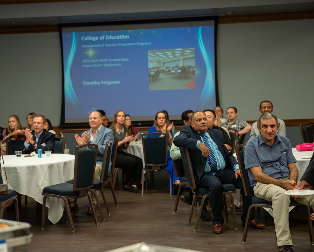 A group of people seated at round tables at the Cohen Student Union. There is a PowerPoint presentation running in the background with photos and names of individuals.  Some individuals in the audience are clapping.  