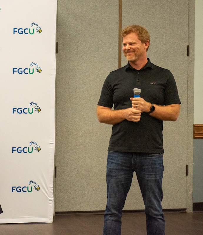 A person stands holding a microphone in front of a banner featuring repeated 'FGCU' logos. They are wearing a black short-sleeve polo shirt, blue jeans, and a black wristwatch. 
