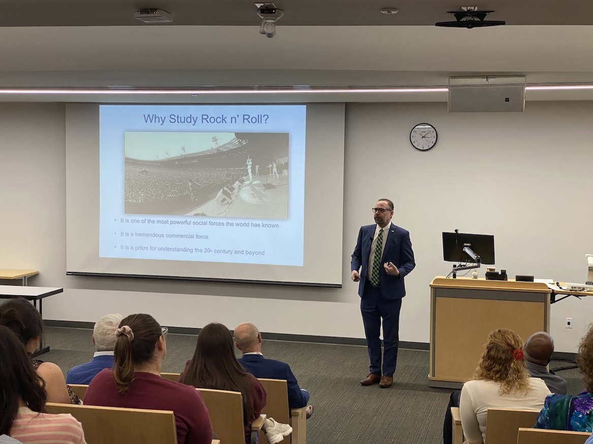 Standing at the front of an auditorium, facing the crowd, Dr. Clay Motley, dean of the College of Arts & Sciences, presents “The Origins of Rock and Roll” as part of FGCU’s “Provost’s Seminar Series.” Dr. Motley is wearing a navy blue suit, white dress shirt and green and black tie.  