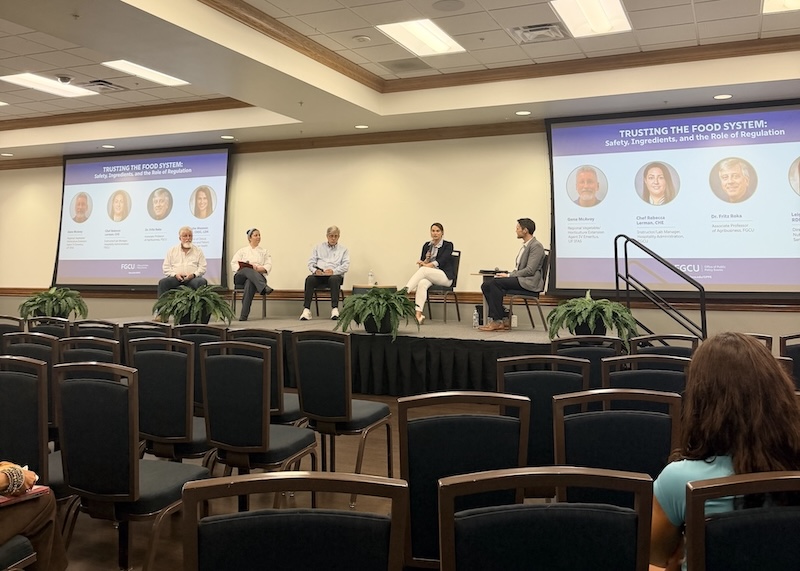 Five panelists sit on a stage in a conference room during a discussion. Two projection screens display the event title, “Trusting the Food System: Safety, Ingredients, and the Role of Regulation,” while audience seating is visible in the foreground.