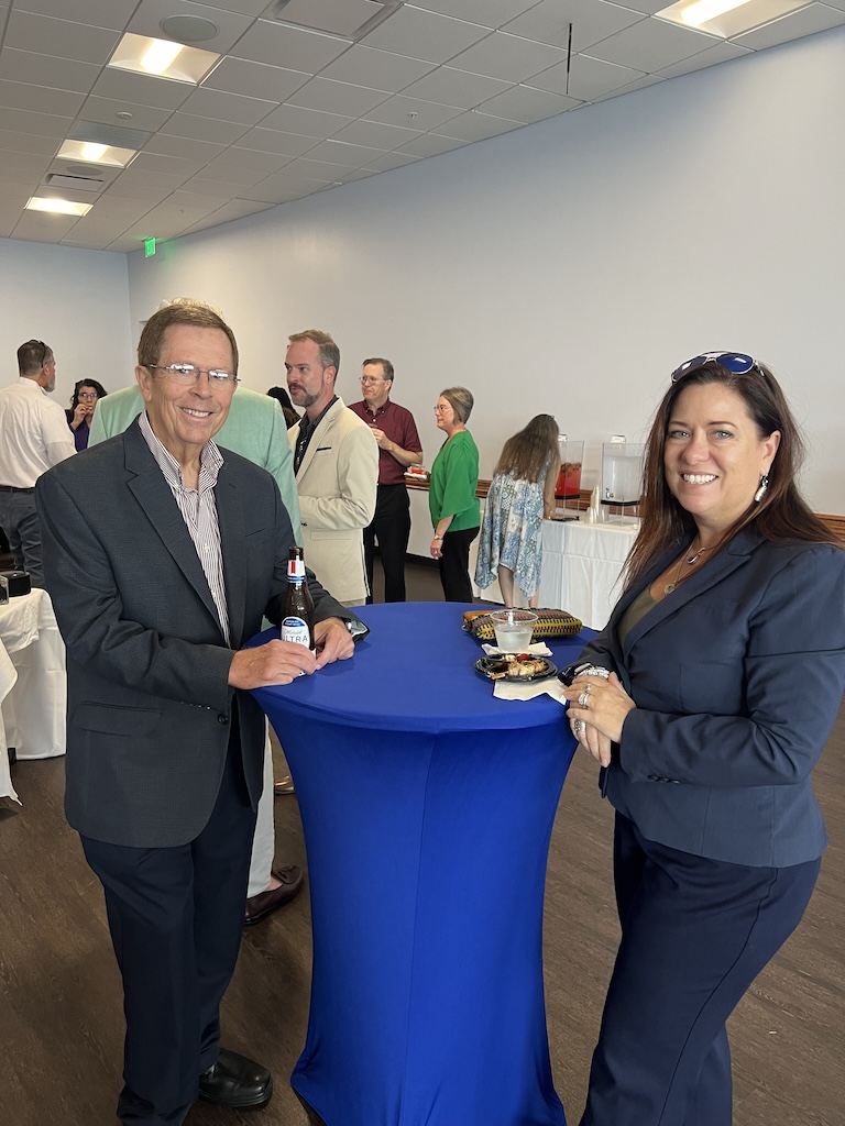 Dr. Mathew Sheep and Andi Clemons pose for a photo as they enjoy refreshments and hors d'oeuvres. Dr. Mathew Sheep is wearing a dark blue blazer, blue pants and a light red dress shirt. Andi Clemons is wearing a navy blazer and sunglasses. People can be seen in the background socializing and grabbing a drink from the beverage table. The beverage table has two beverage containers.  