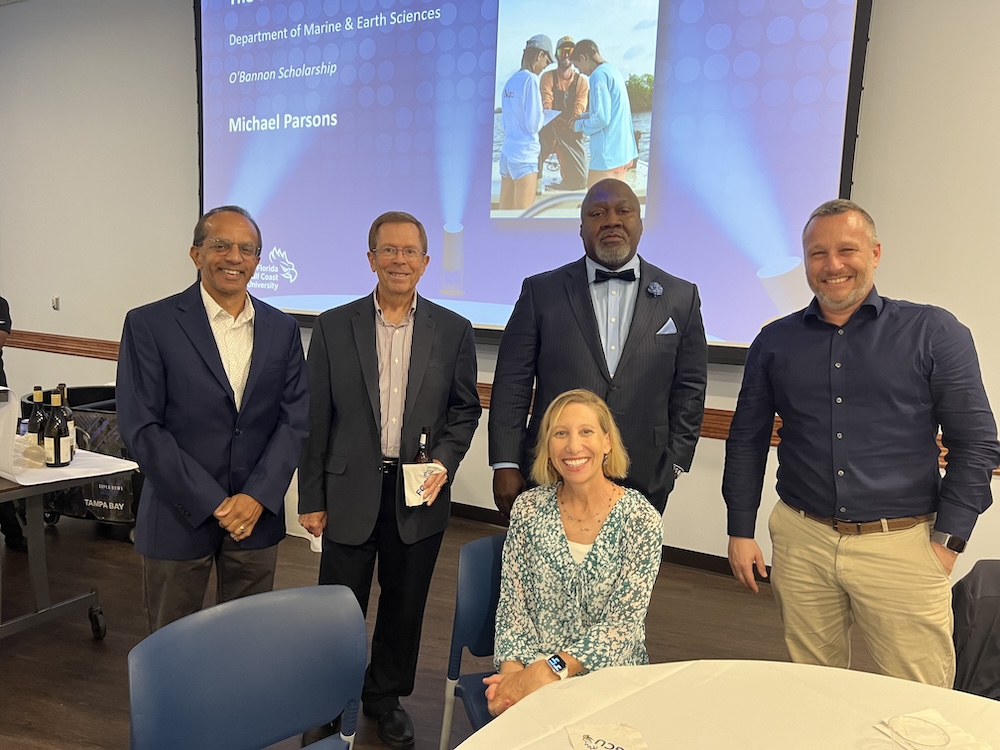 Standing behind a white wall and a presentation screen, four people, and one person sitting down, are posing for a photo. From left to right, Thomas Mathew is wearing a blue blazer, gray pants and a white dress shirt. Dr. Mathew Sheep is wearing a dark blue blazer, blue pants and a light red dress shirt. Interim Provost Tony Barringer is wearing a black suit, light blue dress shirt, black bow tie, light blue handkerchief and a light blue flower pin on his jacket. Dr. Alexander Magnier is wearing a dark blue dress shirt and khaki pants. Sitting down, Dr. Weiss is wearing a green and white blouse, a necklace and watch. A table and blue chair in seen in front of the group.  