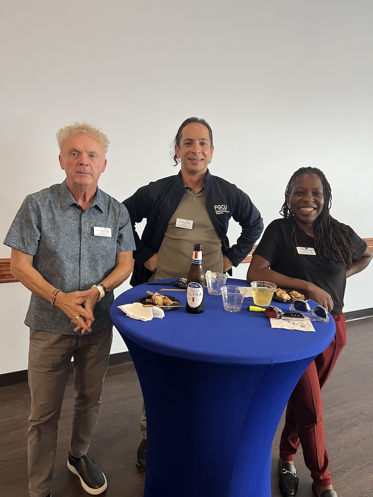 Simon Wood, Ersun Uluc and Charlene Mitchell pose for a photo while enjoying refreshments and hors d'oeuvres at a cocktail table. Simon Wood is wearing a blue button-down shirt and his nametag. Ersun Uluc is wearing a black FGCU zip-up, an olive-green polo shirt and his nametag. Charlene Mitchell is wearing a black polo shirt and her name tag.  