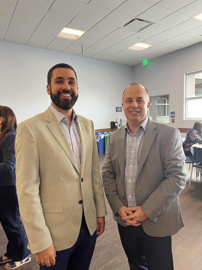 Wearing a cream blazer, black pants and stripped dress shirt, Dr. Amir B. Ferreira Neto is standing next to Dr. Tom Felke who is wearing a gray blazer, gray dress shirt and black pants. A person can be seen standing in the background and a person sitting down by a table. 