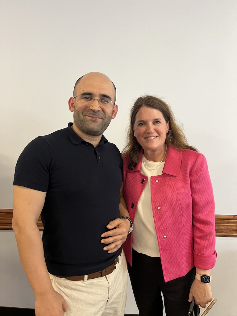In front of a white wall, Dr. Burhan Ozfidan and Dean Diana Chesire are posing for a photo. Dr. Burhan is wearing a black polo shirt and reading glasses. Dean Diana is wearing a pearl necklace, a pink jacket and a beige blouse. 