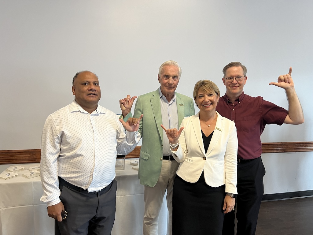  Standing behind a white wall, four people are posing for a photo as they give the Wings Up salute. From left to right, Dean Huzefa Kagdi is wearing a white dress shirt and gray pants. FGCU Board of Trustee Vice Chair Dick Eide, Jr. is wearing a pastel green blazer, light blue dress shirt and khaki pants. President Aysegul Timur is wearing a black dress and a white blazer with a gold FGCU pin. Dr. Glen. Whitehouse is wearing a maroon button-down shirt and black pants. 