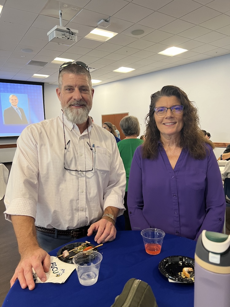 Posing for a photo, Bill Reynolds is wearing a white button-down shirt and black pants; and Dr. Laura Frost is wearing a purple blouse. People can be seen in the background.  
