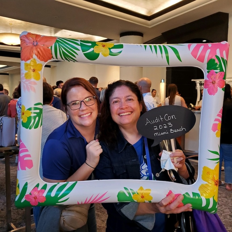 Internal Auditors III Jena Valerioti (left) and Viviana Lauke (right) attend AuditCon 2023 in Miami Beach. Viviana is holding a sign that says “AuditCon 2023 Miami Beach” while they both take a photo with a Hawaiian themed frame photo prop.  