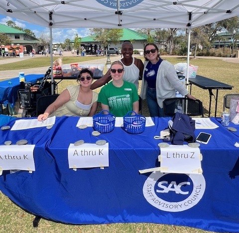Four event volunteers pose behind a blue registration table organized with sign‑in sections under a white canopy outdoors.