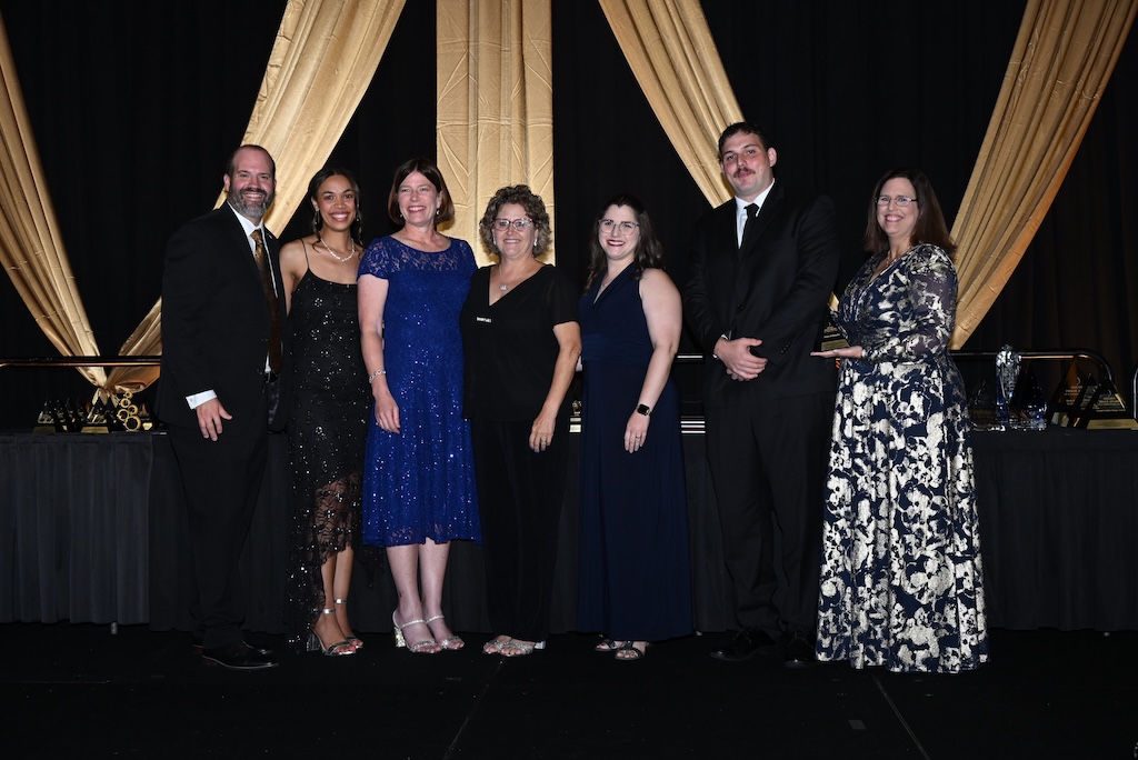 A group of seven individuals dressed in formal attire stand in front of a black and gold backdrop at the Florida Public Relations Association’s Golden Image Awards. 