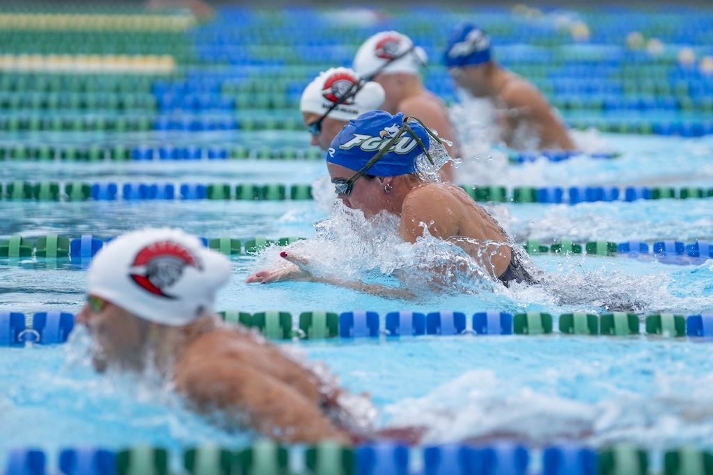 Several swimmers compete in a breaststroke event in an outdoor pool. Each swimmer is in a separate lane marked by alternating green and blue lane dividers. The swimmer in the center wears a blue swim cap with ‘FGCU’ printed in white and green letters, while others wear white caps with red and black logos.