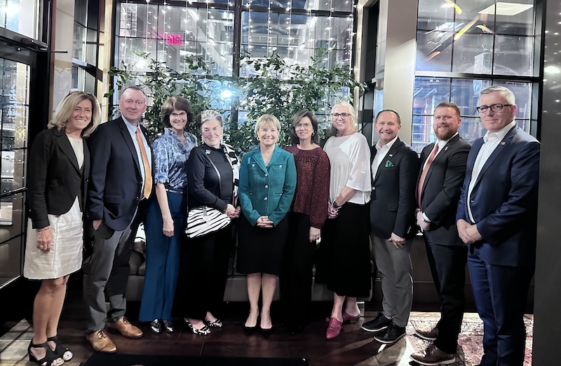 FGCU leadership is posing for a photo. They are wearing professional attire and are in a room at Syracuse University with windows in the background. Buildings can be seen through the windows as well as some greenery.  