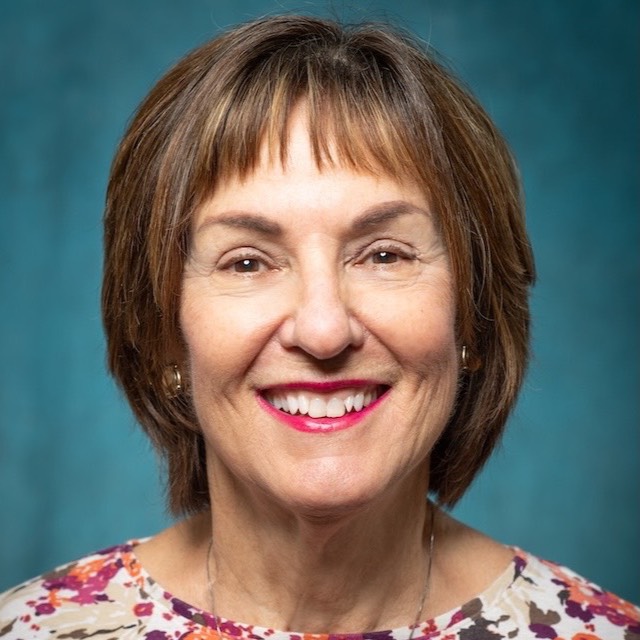 Headshot of Teresa Clark. She has short brown hair and is wearing a necklace and a blouse with an orange, purple, brown and beige flower pattern.