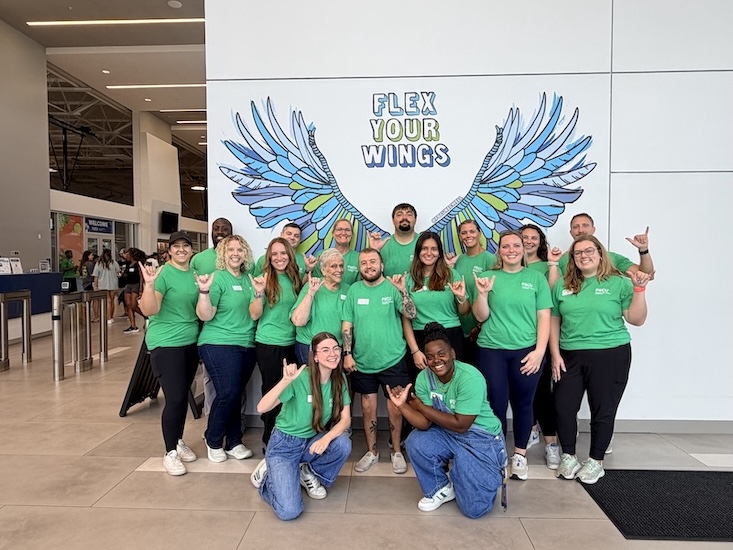 A group of FGCU staff wearing green shirts pose for a photo inside the university recreation building. The group stands in front of a large mural featuring blue and green wings and the words “Flex Your Wings,” with several participants making the FGCU hand sign.