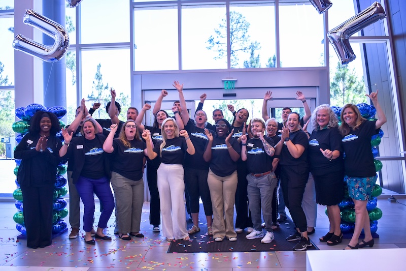 University Recreation & Wellness (URecWell) employees celebrate their one millionth visitor at the URecWell Center. Large windows are in the background.  The group is posing for a photo, with many individuals making celebratory gestures. Green and blue balloon decorations are on both sides of the group, and confetti is on the floor. Employees wear matching black T-shirts.  