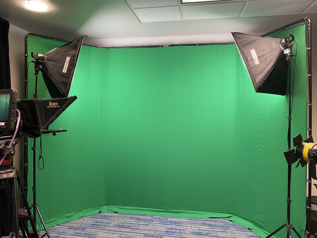 An indoor video studio features a large curved green screen surrounded by professional lighting stands. A camera with a teleprompter is positioned to the left, and additional studio lights are mounted around the backdrop.