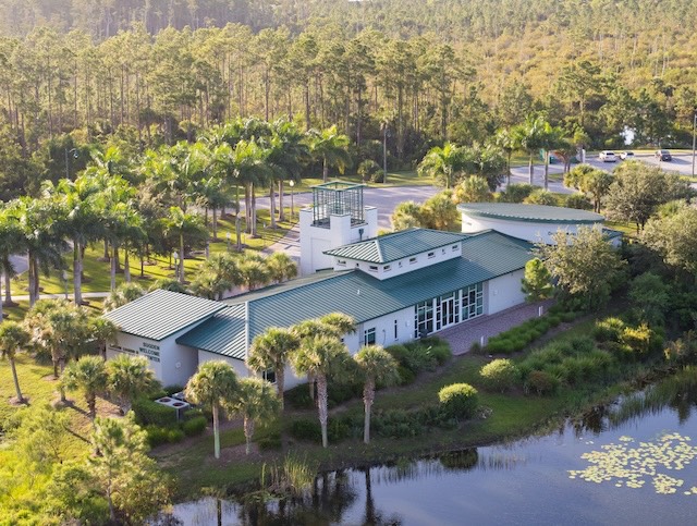 Aerial view of the Margaret S. Sugden Welcome Center on a sunny day. The welcome center is surrounded by greenery, palm trees, a forest to the north side of the center and a lake on the south side. 