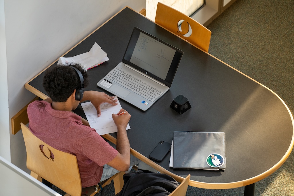 Aerial view of a male wearing a red shirt and headphones sitting down on a chair while he is writing on a piece of paper that is on a table. A laptop, gray folder with an Ģ��TV eagle sticker on the front, and a cell phone are on the table.