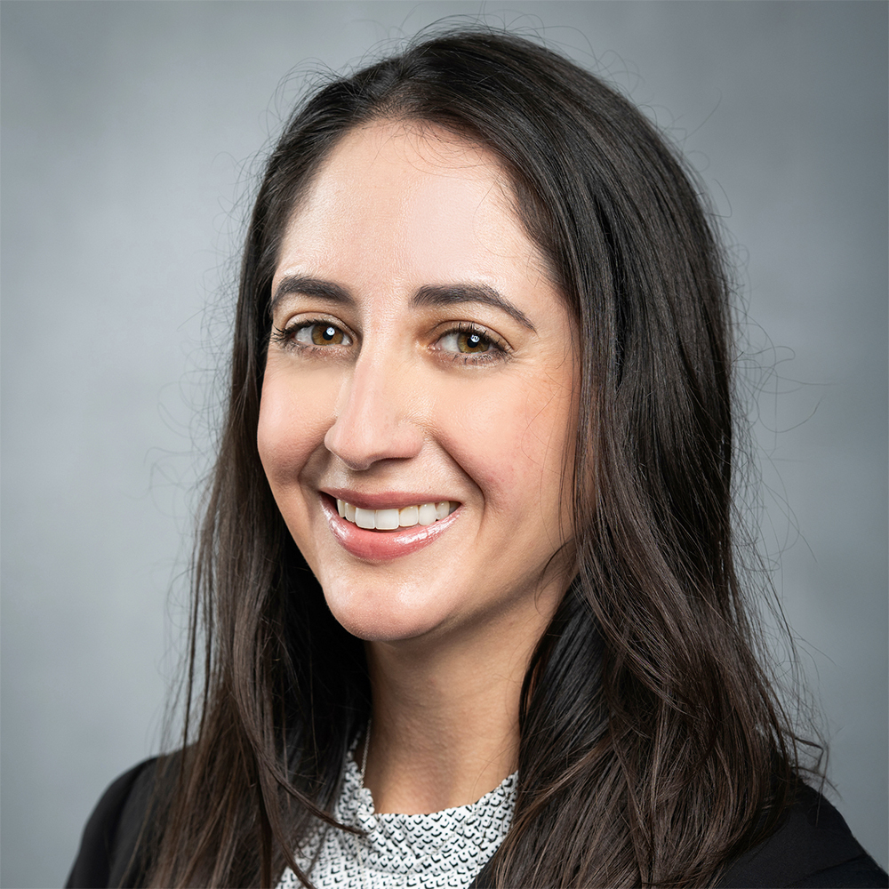 A person with long dark hair is dressed in a black blazer layered over a light top, standing against a plain gray background.