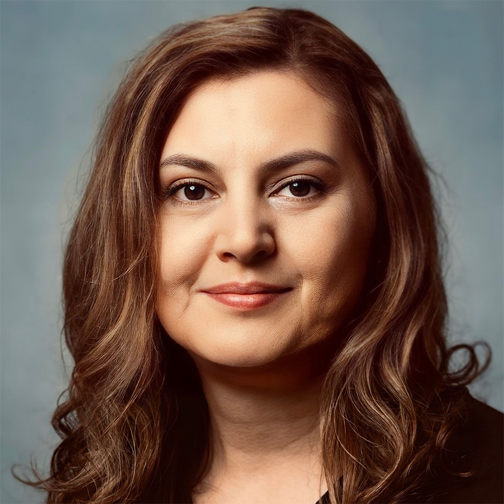 A person with long, wavy brown hair stands in front of a neutral, plain background.