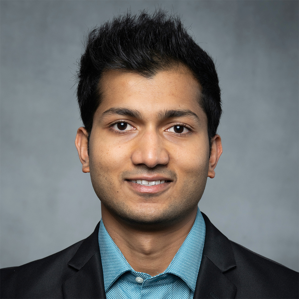 A man with short dark hair, dressed in a blue dress shirt and black blazer, standing against a plain gray background.