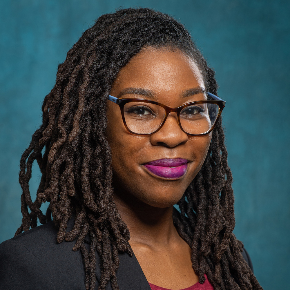 A female with long, twisted hair and glasses is dressed in a dark blazer layered over a red top. The background is a smooth, solid teal color.